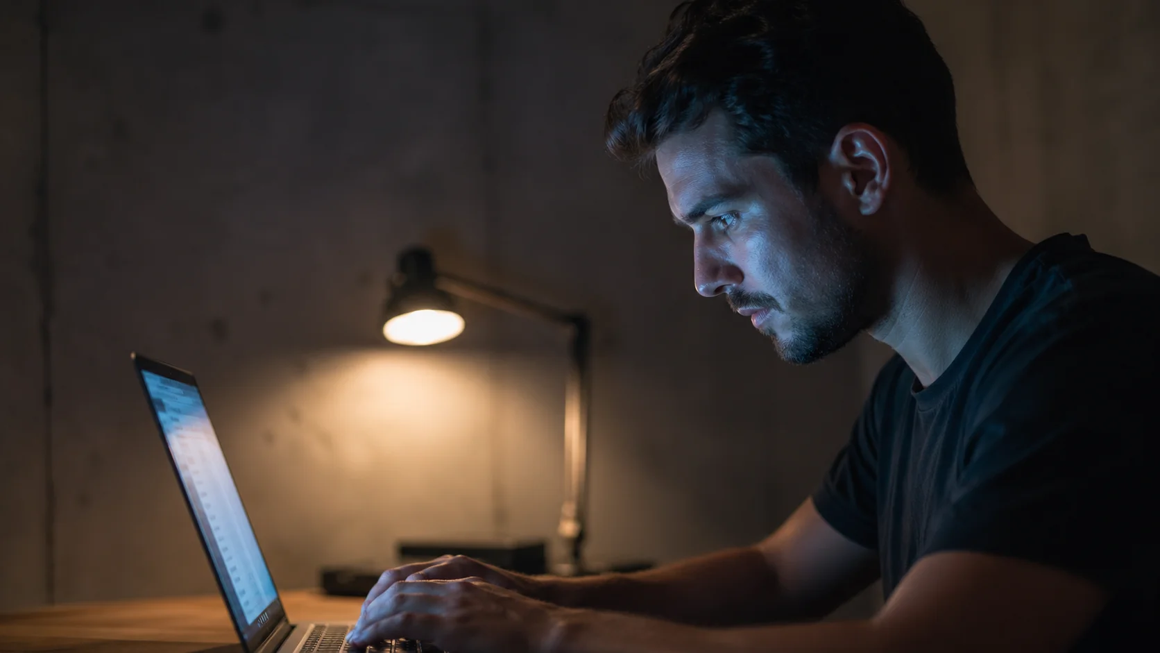 Close-up side profile of a man typing on a laptop in a dimly lit room with the screen casting a blue glow on his face